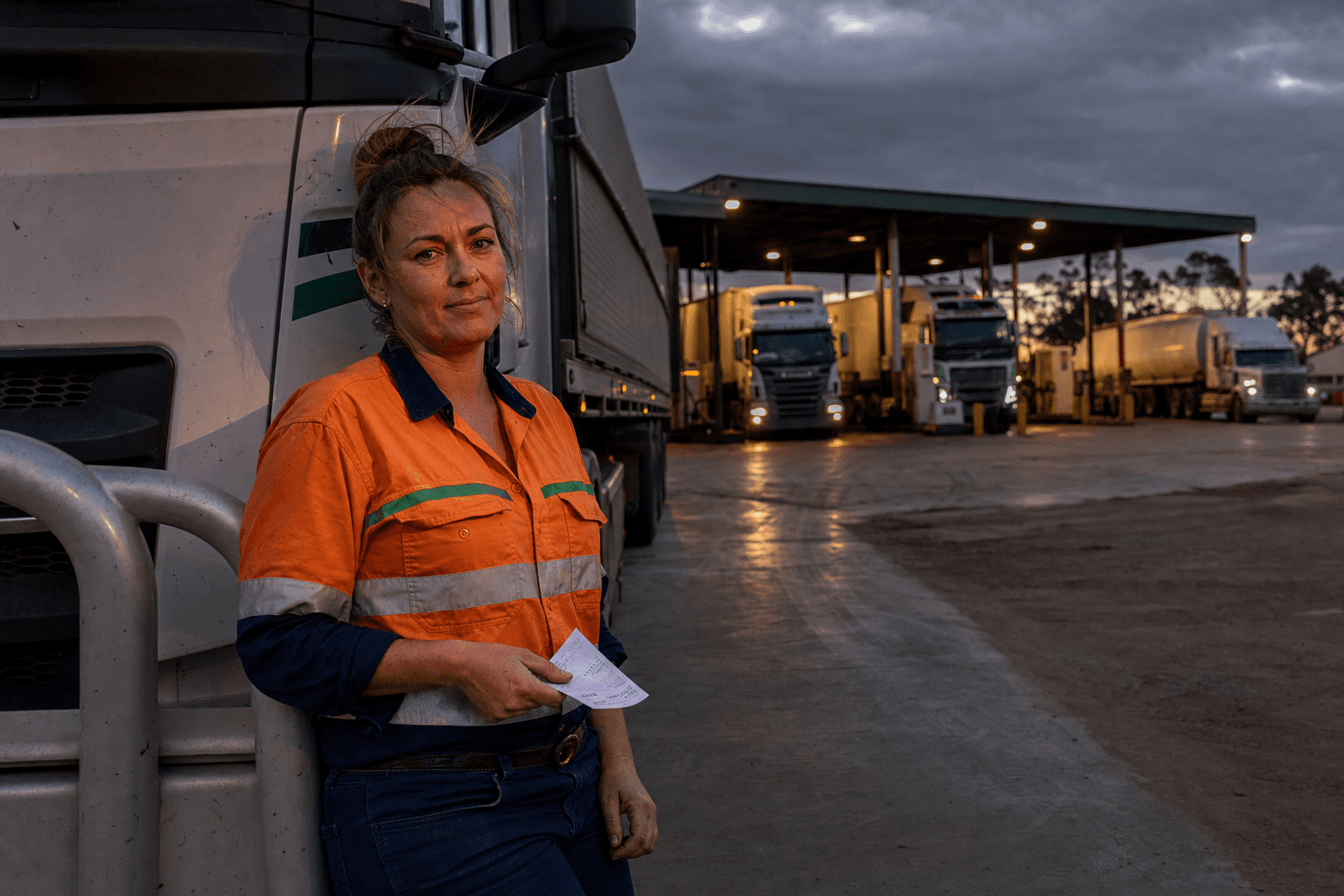 A female truck driver in orange high-vis leans against a B-double cab at a regional Australian fuel stop at dusk, holding a fuel receipt, with other trucks queuing at diesel bowsers behind her.