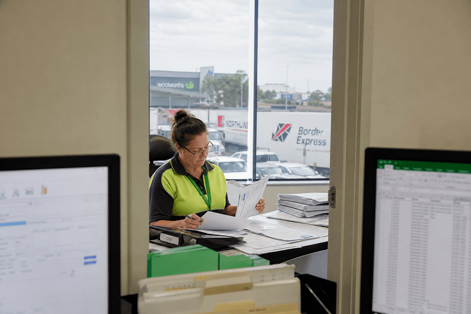 Seen through a doorway between two monitors, an office worker manually checks paper invoices and packing lists against a Certificate of Origin form in a bright, daylit freight office.