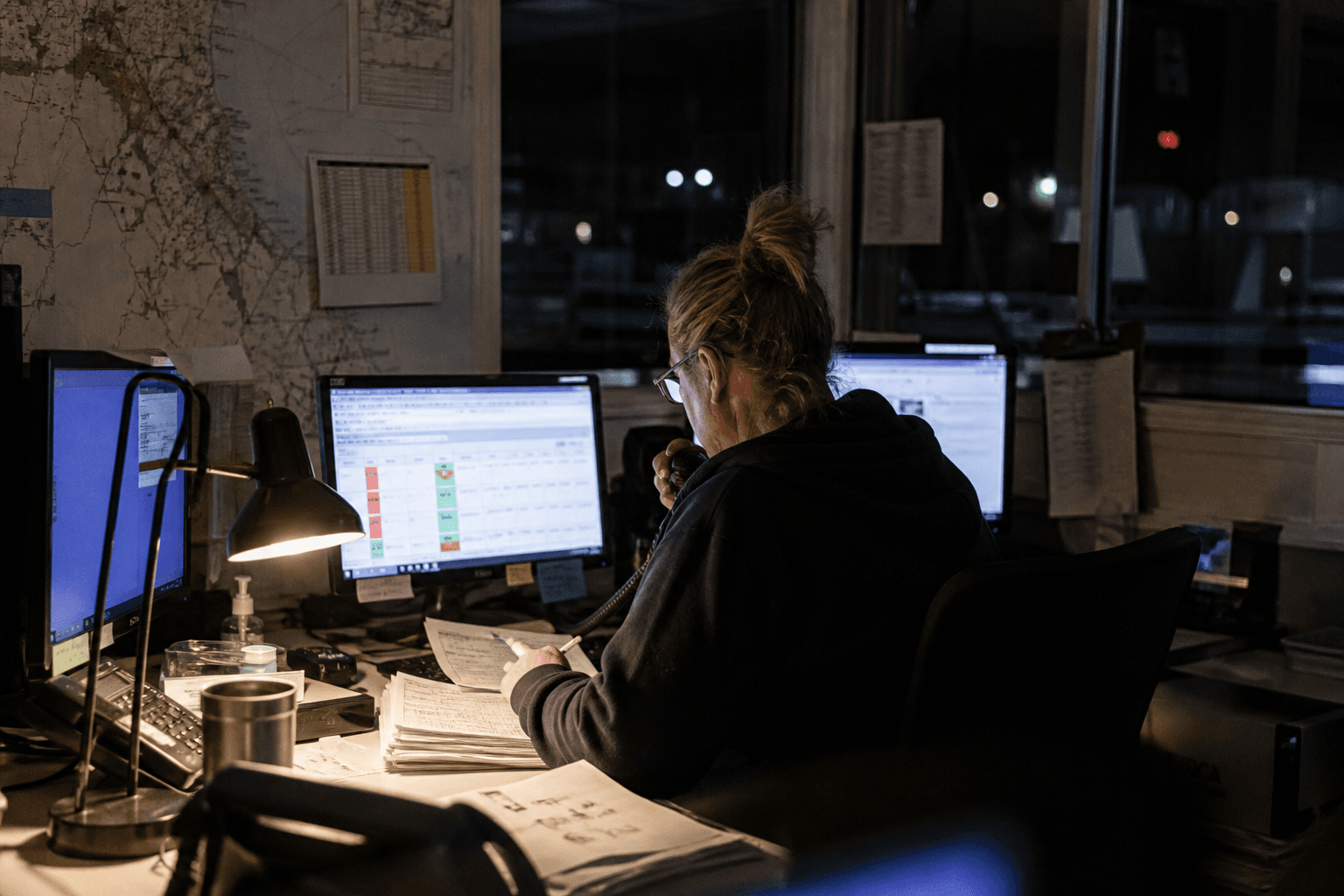 A dispatch coordinator sits at a night-shift desk in a freight depot office, lit by a desk lamp and computer screens showing scheduling software, with route maps on the wall behind them.