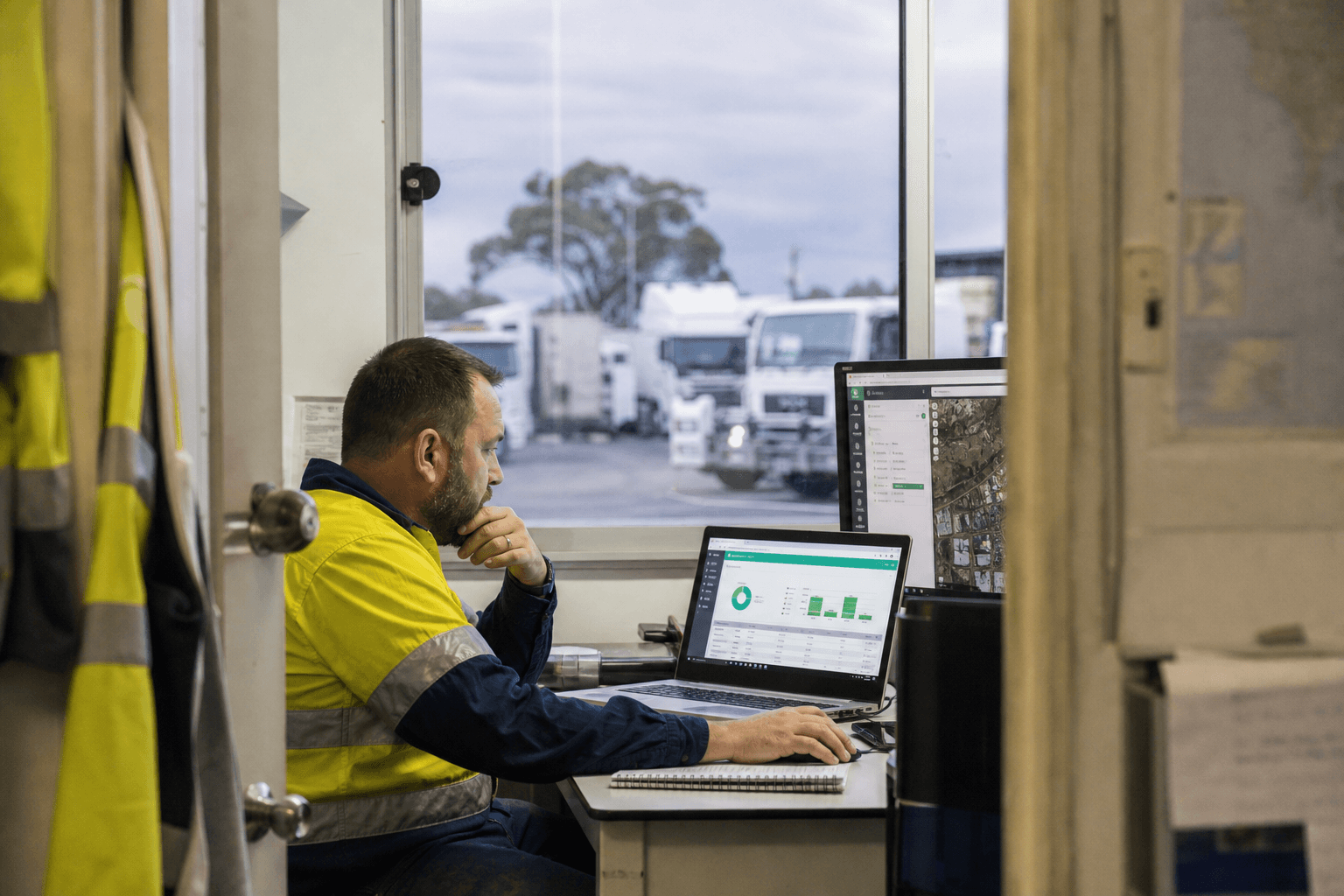 An operations coordinator reviews a fleet data dashboard on a laptop in a depot office, framed through a doorway with a truck yard visible through a window behind them.