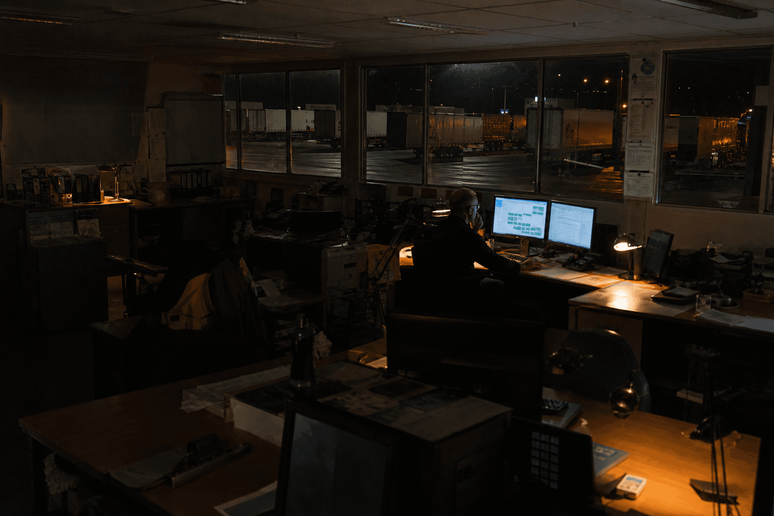 A dispatcher works alone at a desk with glowing dual monitors showing a load plan, inside a dimly lit freight dispatch office at night with a dark trailer yard visible through windows.