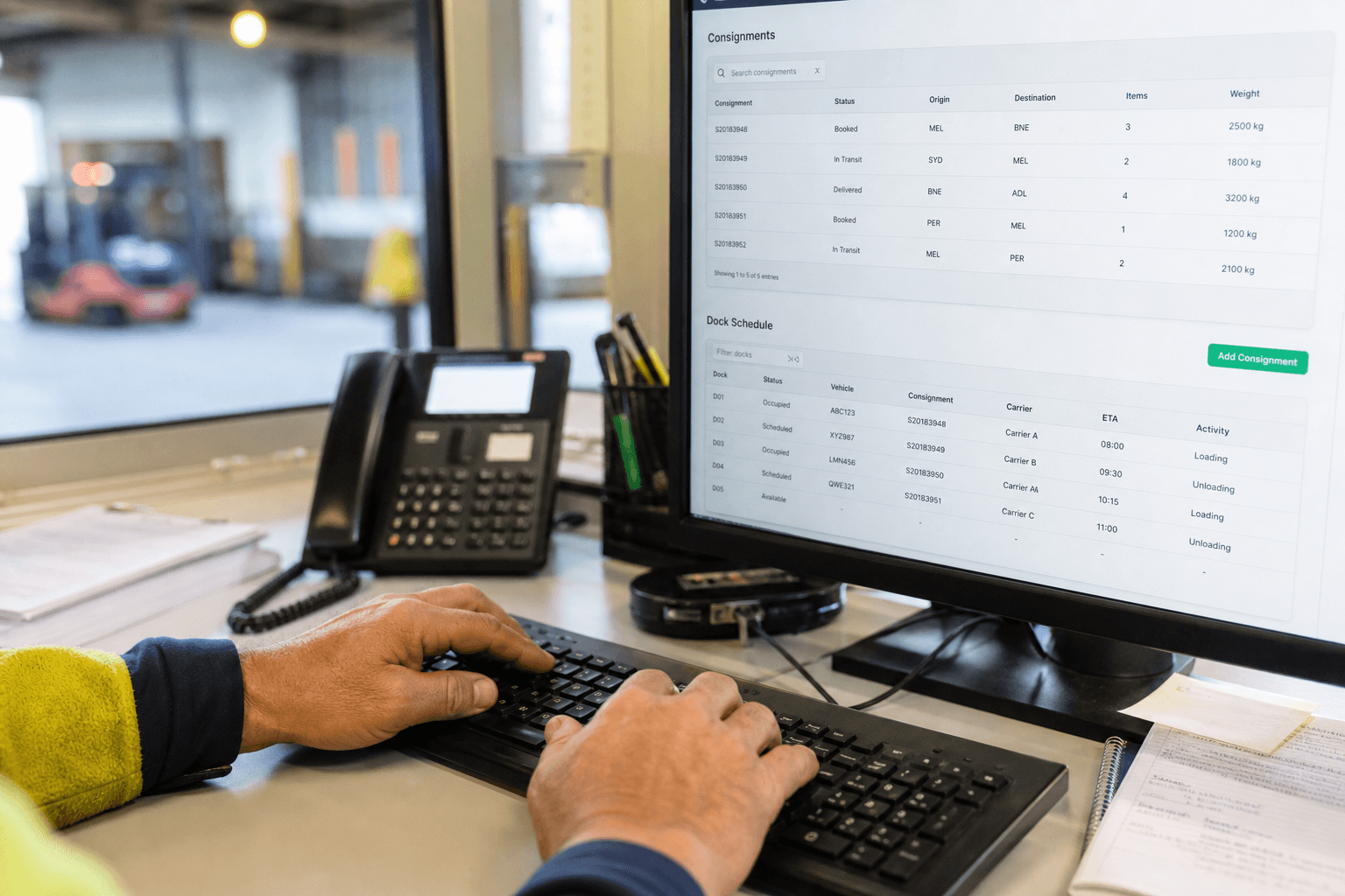 Close-up of a person's hands typing on a keyboard with a transport management system screen showing shipment and dock data visible in the background.