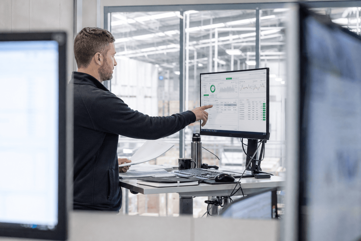 A logistics coordinator viewed between two monitors, pointing at an automated performance dashboard on screen in a bright, daylit depot office with a warehouse visible behind glass.