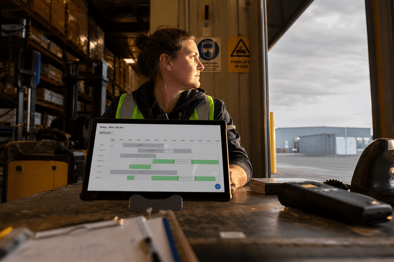 Low-angle view of a dispatcher checking a digital rostering screen in a warehouse, lit by golden afternoon light from an open dock door.