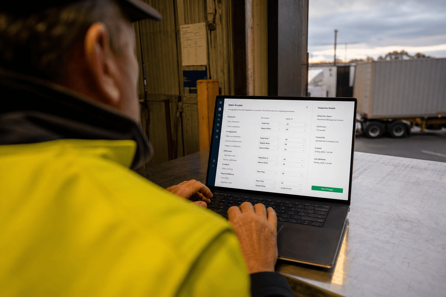 Over-the-shoulder view of a logistics operations worker reviewing data access settings on a laptop at a dock bench during late-afternoon light.