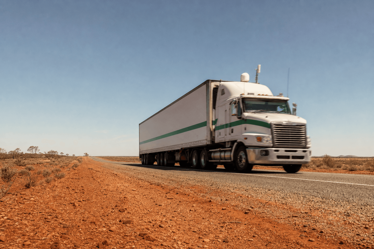A refrigerated semi-trailer travels along a remote outback Australian highway under a broad pale sky, with a satellite antenna dome and cellular antenna visible on the cab roof, surrounded by flat red-dirt scrubland stretching to the horizon.