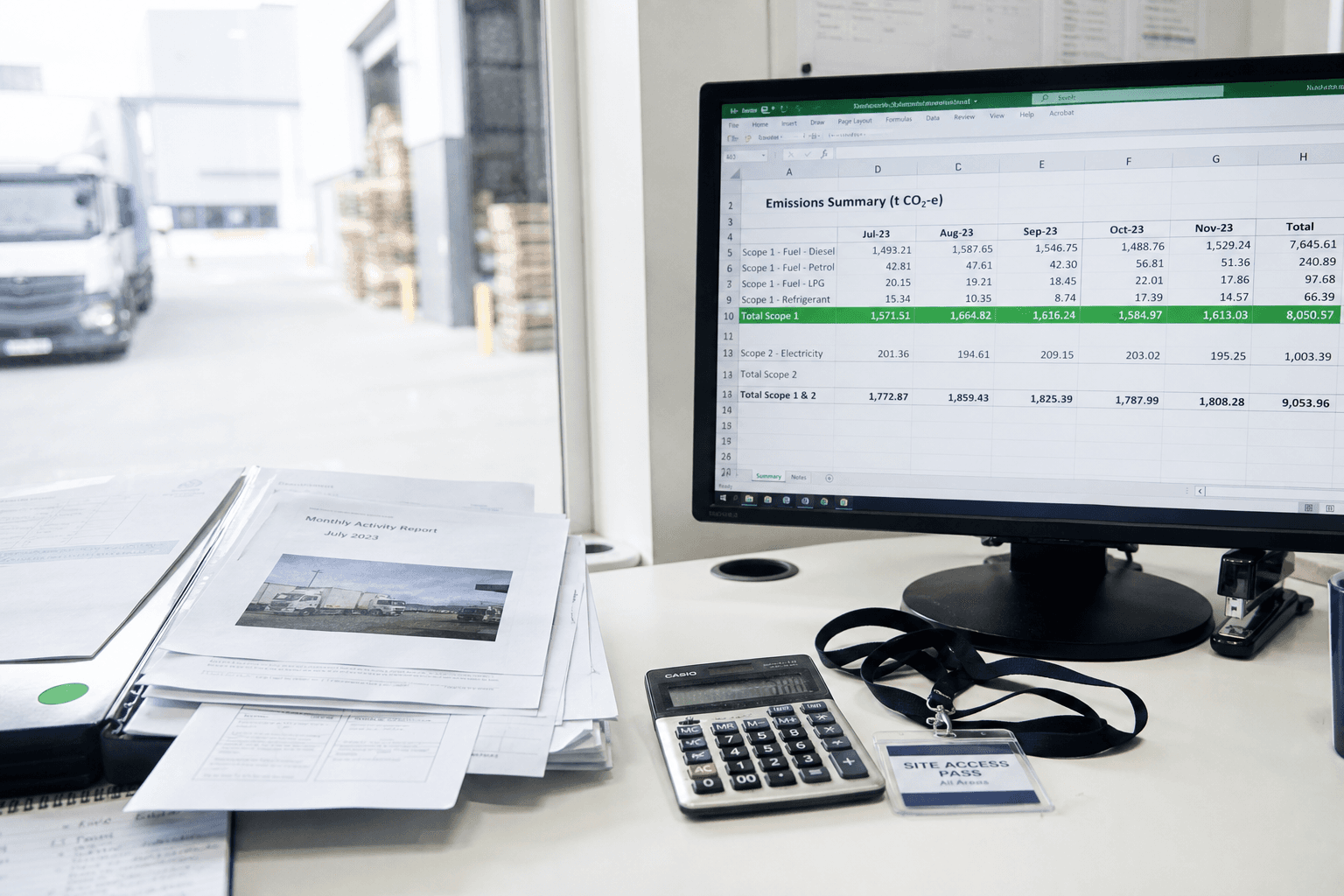 A bright office desk showing a monitor with an emissions data spreadsheet next to printed activity reports and a calculator, lit by natural daylight from a window.