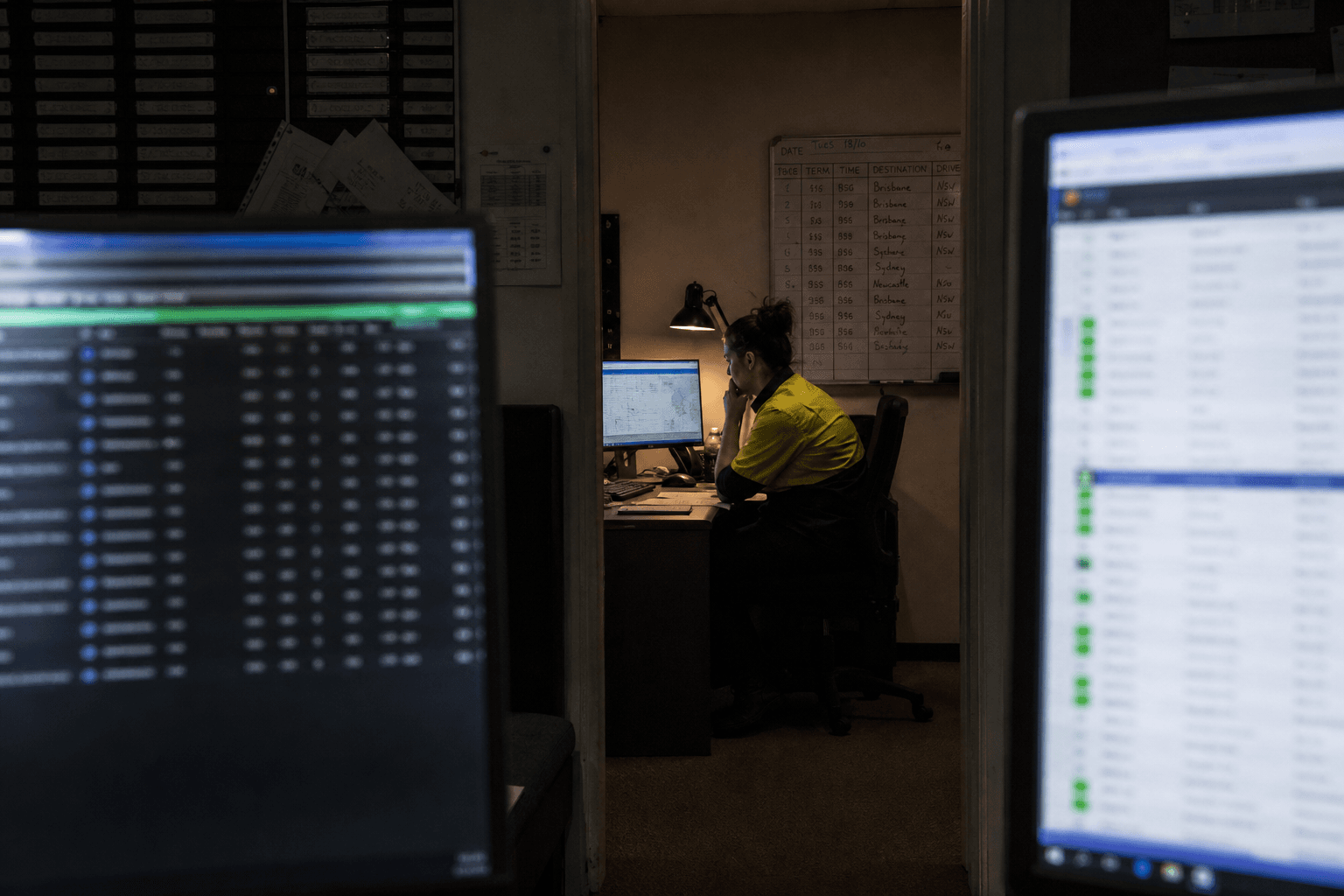A dispatch coordinator viewed through a doorway between two monitors works late in a dimly lit freight control room, illuminated mainly by screen glow and a desk lamp.