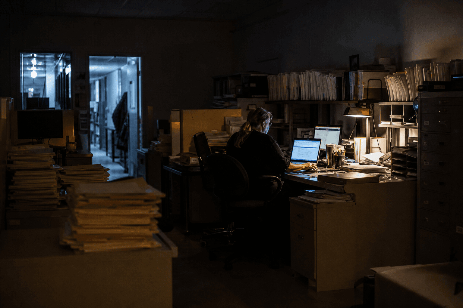 A compliance worker sits alone at a desk in a dim office at night, dwarfed by rows of filing cabinets, lit by screen glow and a desk lamp.