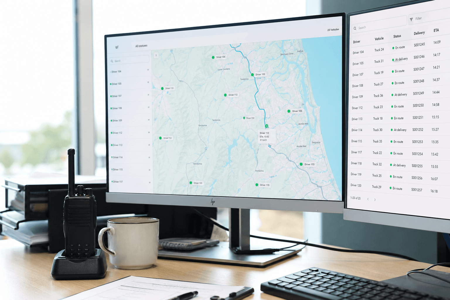 A dispatch desk with a monitor showing a fleet tracking map and GPS markers, a data screen, a radio, and a coffee cup, lit by bright daylight, with no person in frame.