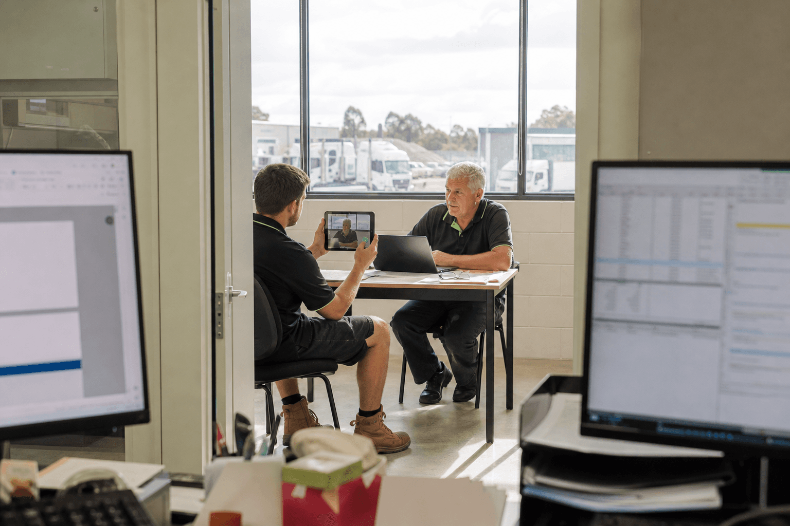 View through an office doorway, framed by monitors, of a younger staff member holding a tablet while interviewing an older operations manager at a table in a sunlit depot meeting room.